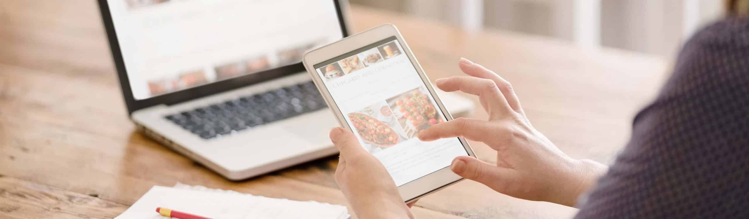 Cropped shot of the rear view of a female blogger reading information from social media holding digital tablet, with book and laptop in table.